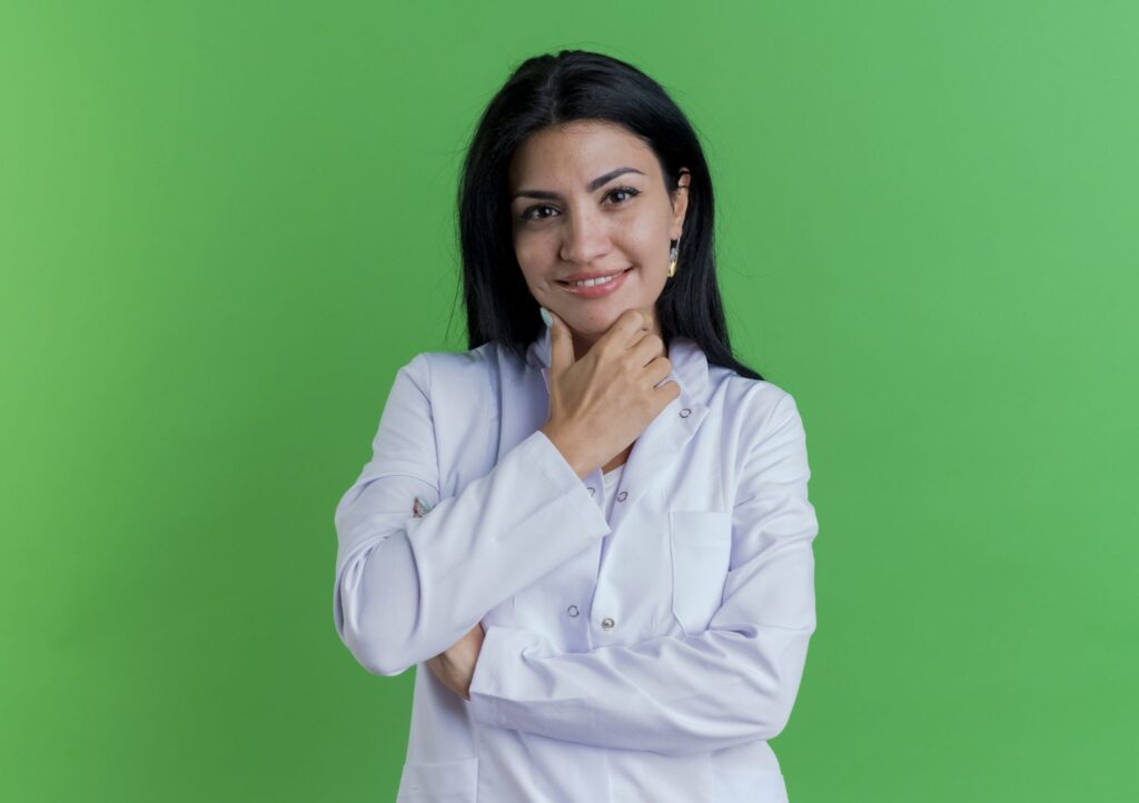 Smiling young female doctor wearing medical robe touching chin isolated green wall with copy space scaled