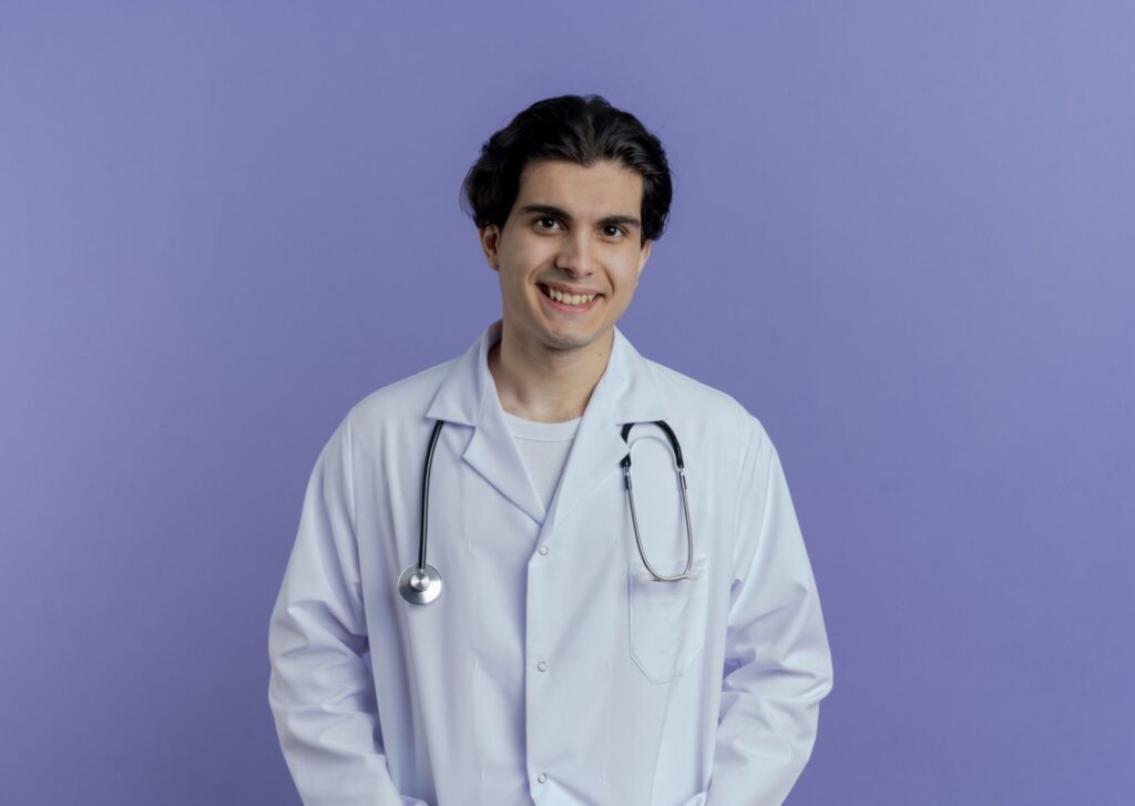 Smiling young male doctor wearing medical robe stethoscope standing isolated purple wall with copy space scaled